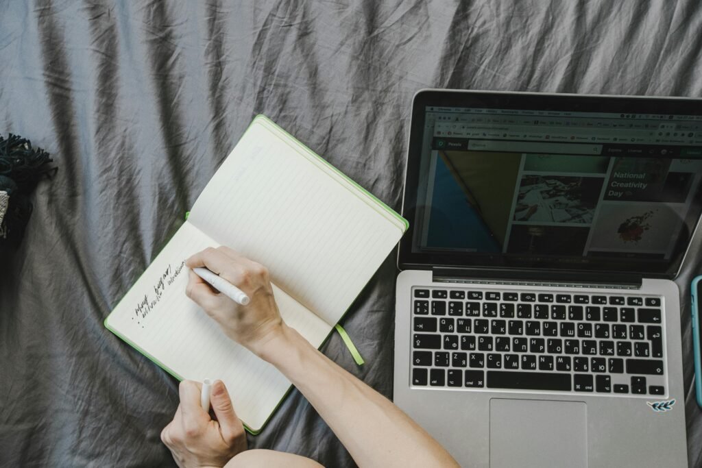 Woman writing in a notebook beside a laptop on a bed, depicting creative freelance work.